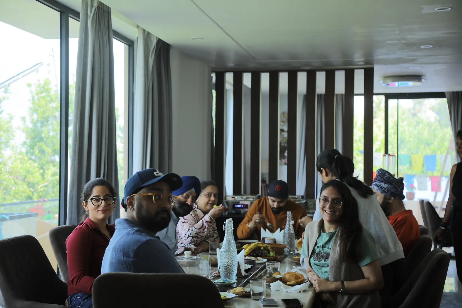 Guests sharing a meal around a long dining table beside tall windows at Cafe DO NTHNG in Manali, several smiling at the camera