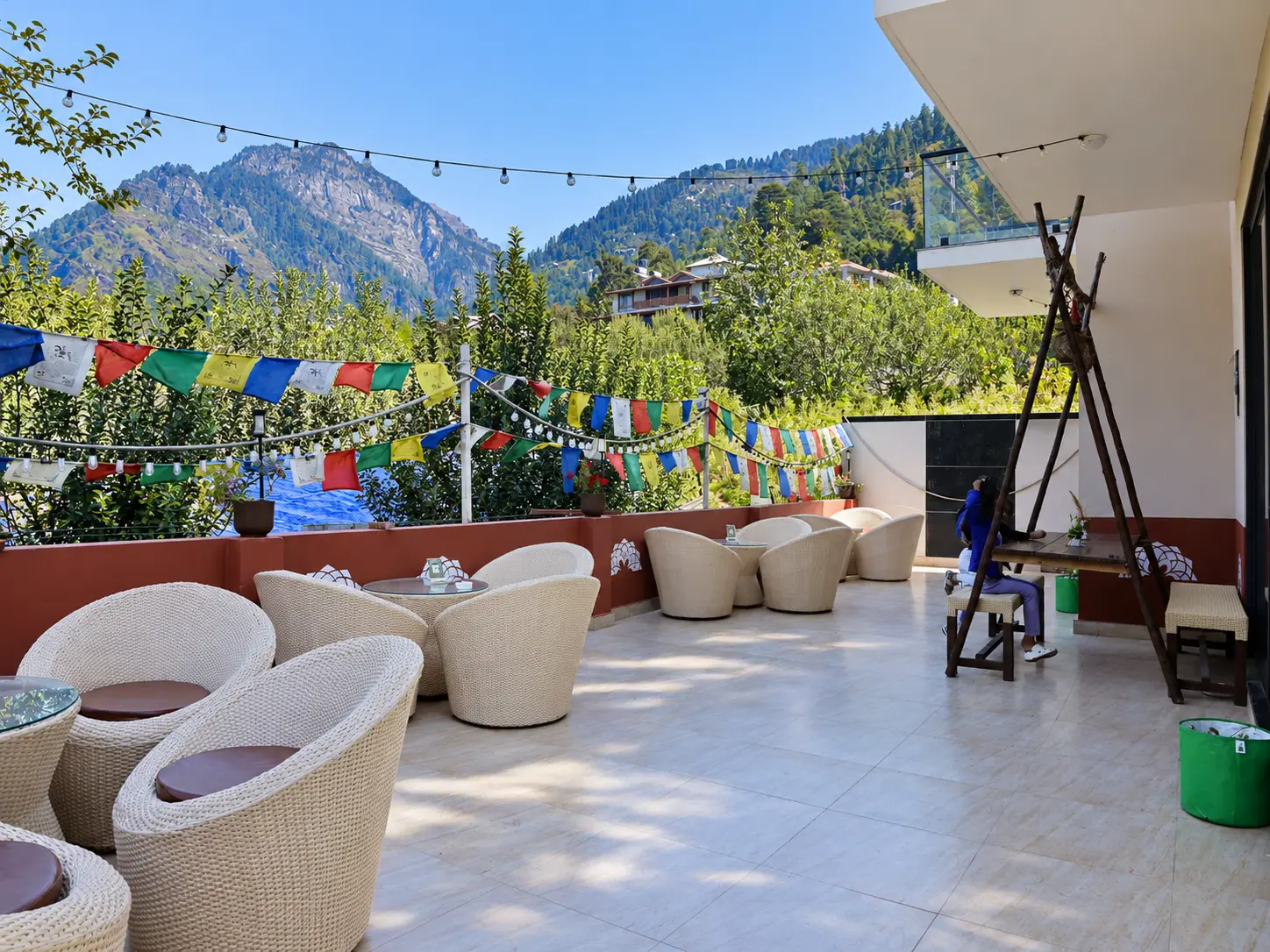 Sunny terrace at Cafe DO NTHNG in Manali, with cream wicker chairs, strings of prayer flags and fairy lights, a wooden swing and forested mountains behind