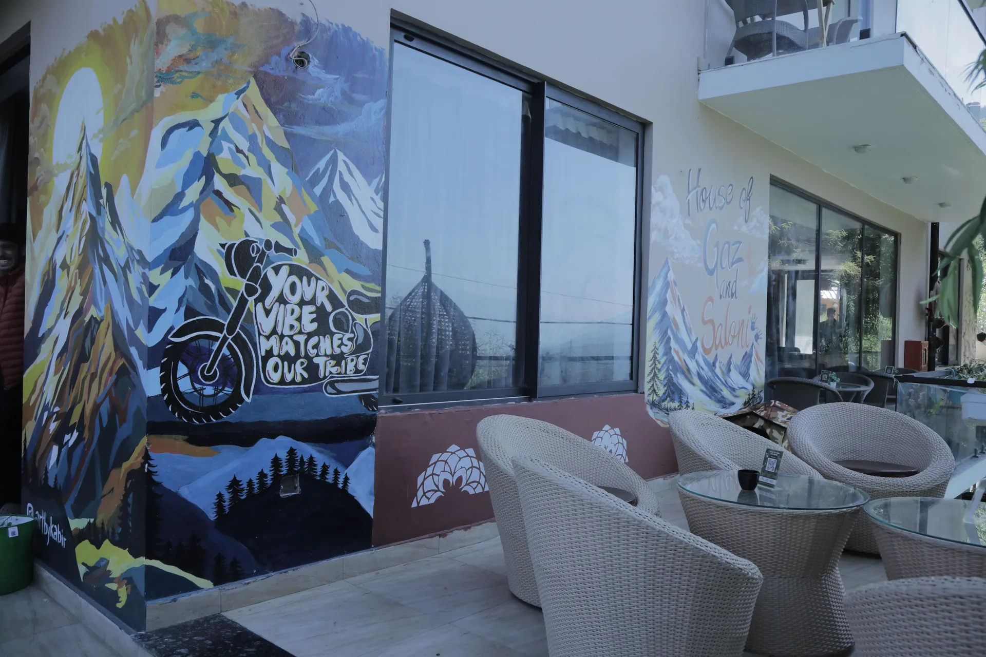 Cafe DO NTHNG terrace in Manali, with a painted mural of a mountain and a motorbike, and wicker chairs around glass tables