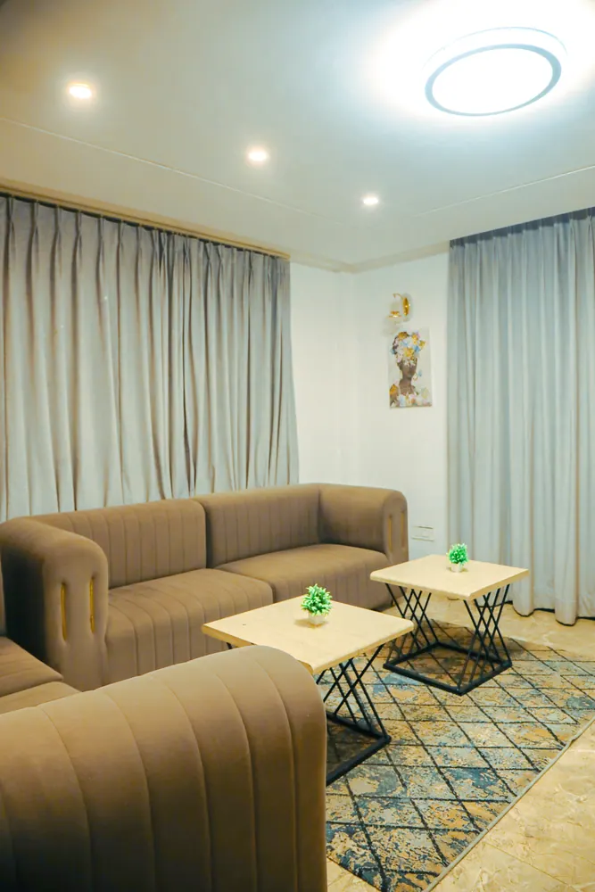Lounge corner with a taupe sofa, two small side tables and a patterned rug beside pale curtains