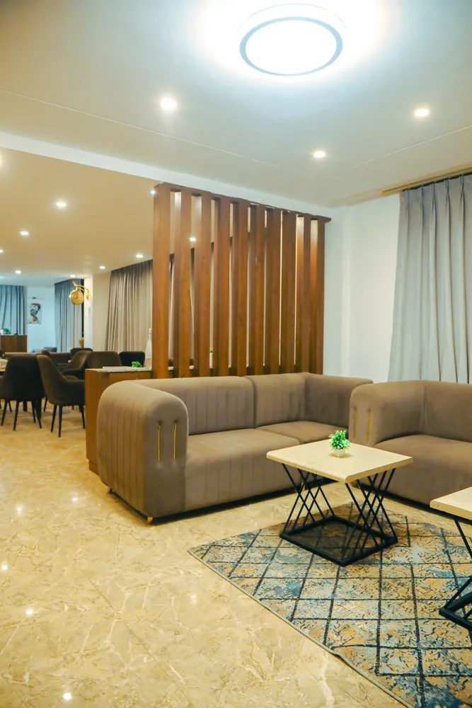 Lounge with a taupe sofa and side table beside a wooden slat partition, with a dining area visible beyond