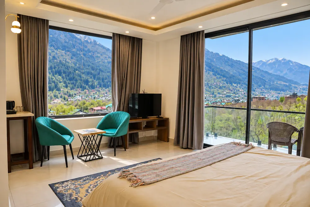 Bedroom at HOGS Panorama, Manali, with teal armchairs and floor-to-ceiling windows overlooking the valley and snow peaks