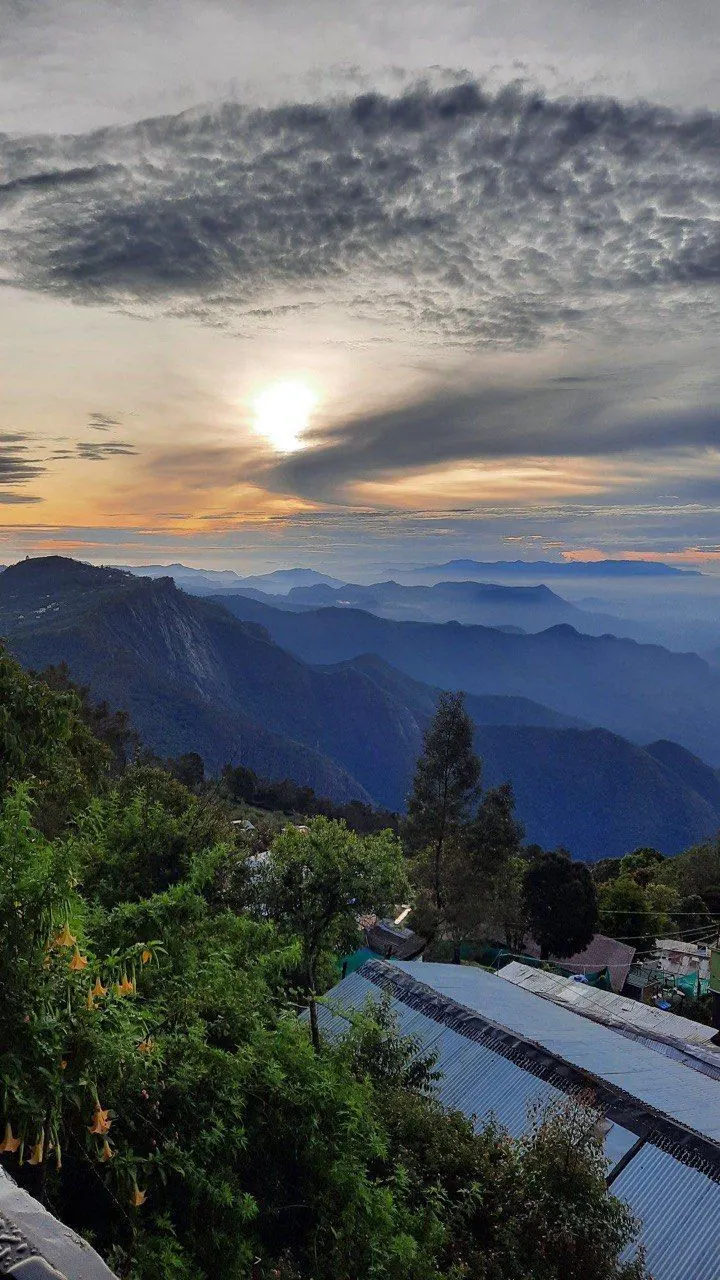 Sunrise glowing through layered clouds over blue mountain ridges, with green trees and tin rooftops in the foreground, seen from HOGS Panorama in Manali